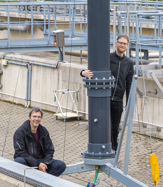 A photography of our team. In the foreground you can see a long, approximately 40 centimeter thick pipe standing on a steel tripod. Our Managing Director André Spindler is sitting on the tripod and smiling into the camera. Our software developer Alexander Walther looks out from behind the pipe. In the background you can see a tank of a sewage treatment plant filled with brown sludge.